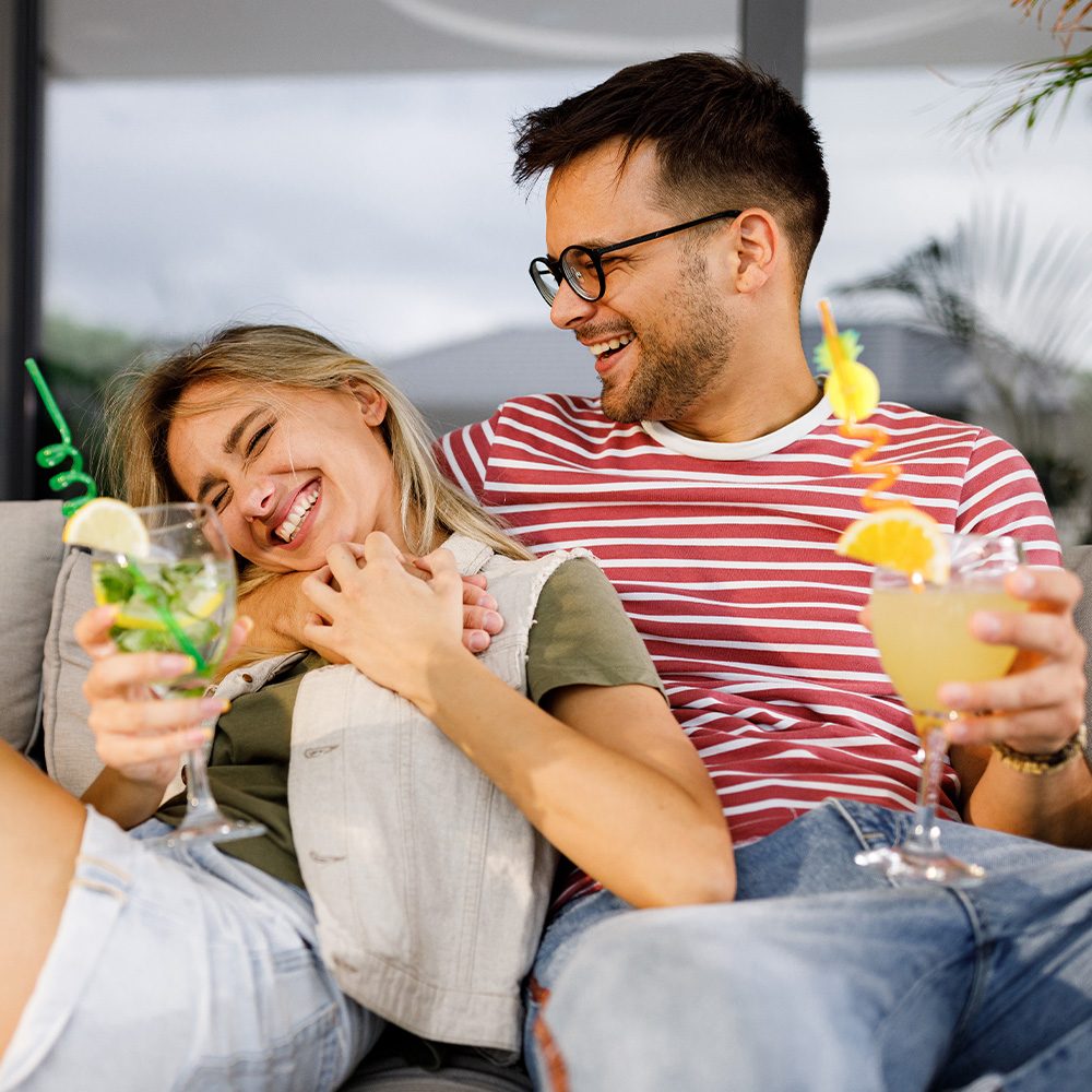 Man and woman sitting on couch laughing while each holding a drink in hand.
