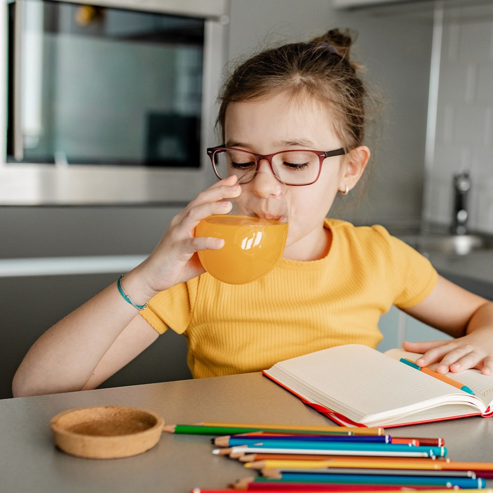 Young girl sitting and drinking orange juice with a book and pencils spread on counter.
